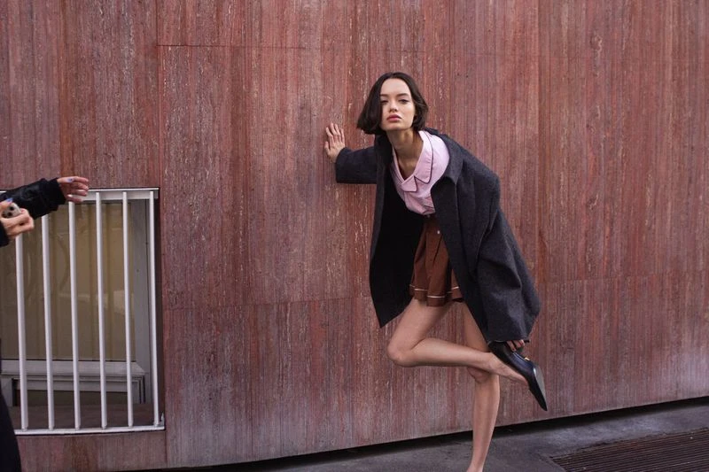 Brunette fashion model strikes a pose in an urban setting with a pink shirt, brown skirt, and grey coat against a textured red wall.