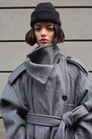 Model wearing an oversized grey pinstripe coat with an exaggerated high collar and a black beanie against a grooved stone wall.