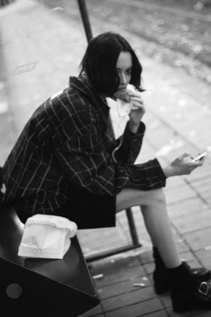 Model in a plaid jacket sitting on a bench eating a snack and holding a phone in a black and white photograph.