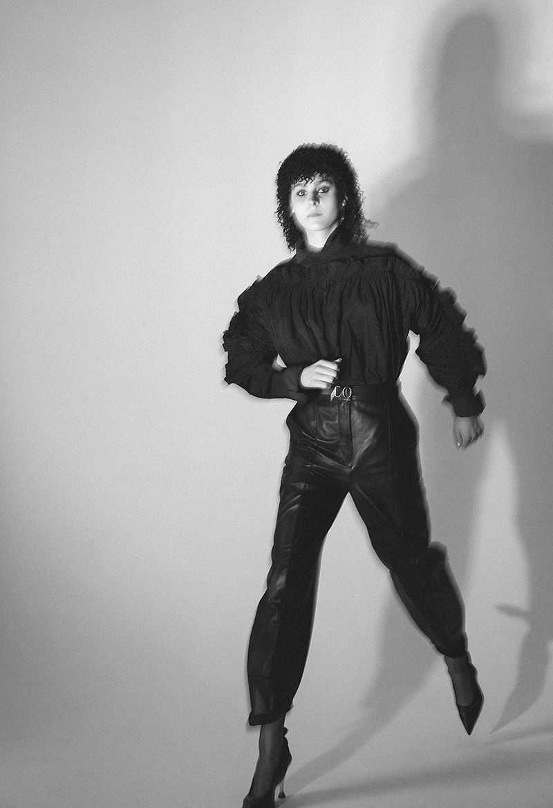 Woman in black blouse and leather pants strikes a dynamic pose in a photo studio, set against a neutral backdrop with her shadow visible.