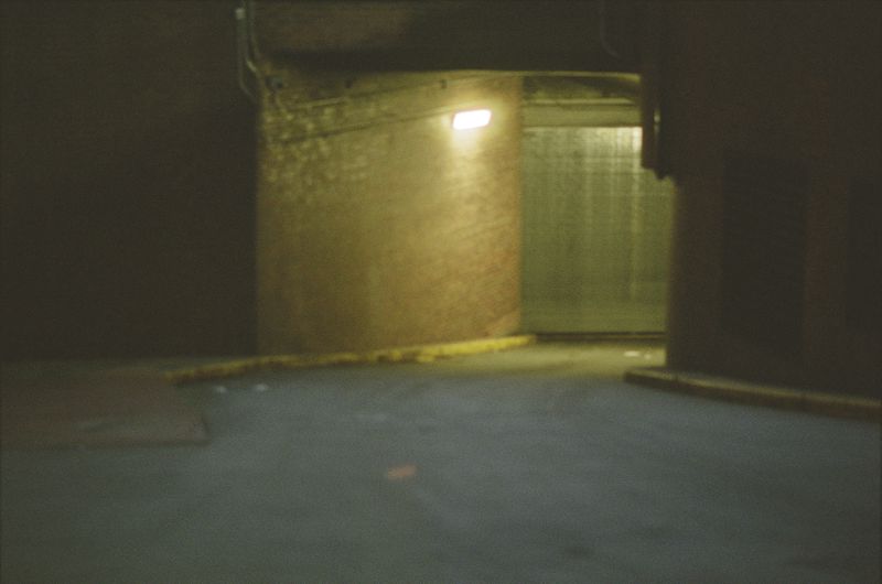 Dimly lit concrete interior with a metallic door at the end of a hallway and a light fixture on the wall.