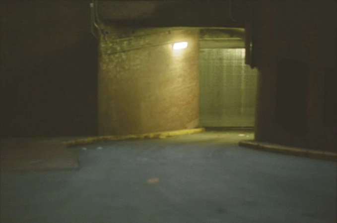 Dimly lit concrete interior with a metallic door at the end of a hallway and a light fixture on the wall.