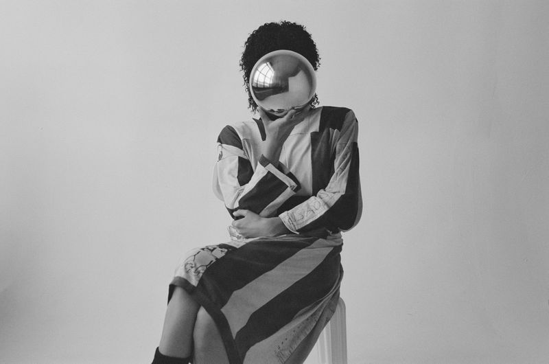 Monochrome image of a person with an afro hairstyle, wearing a striped top and skirt, seated with a silver sphere obscuring their face.