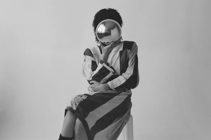 Monochrome image of a person with an afro hairstyle, wearing a striped top and skirt, seated with a silver sphere obscuring their face.