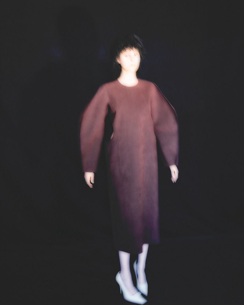 Model in a long burgundy structured-sleeve dress and white high heels posed against a black backdrop in soft focus.