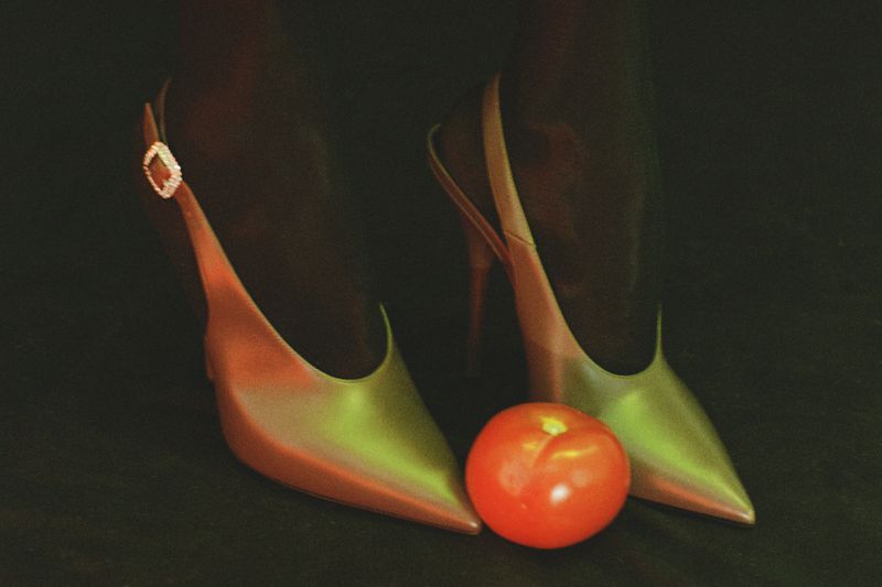 Stylish red and green high-heeled shoes on dark background, complemented by a round, vibrant red tomato placed in front.