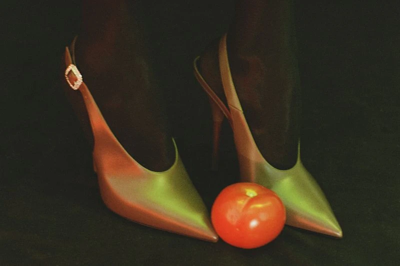 Stylish red and green high-heeled shoes on dark background, complemented by a round, vibrant red tomato placed in front.