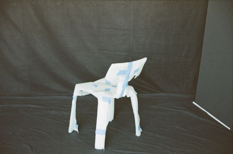 Abstract studio shot of a deconstructed white chair wrapped in blue tape against a black backdrop with a geometric panel on the right.