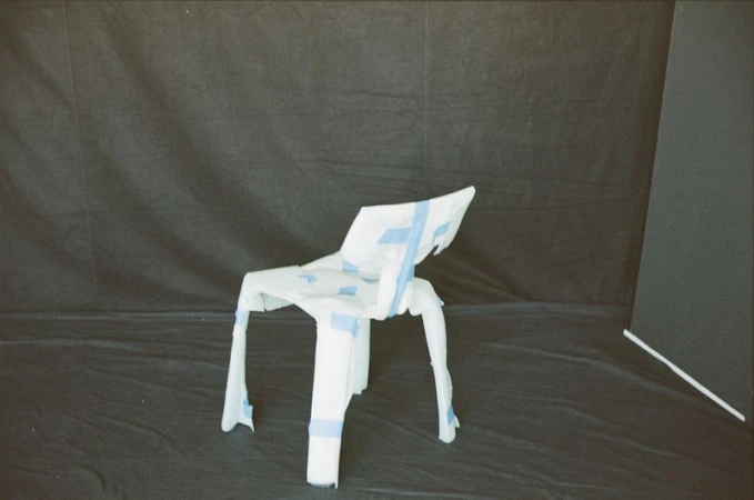 Abstract studio shot of a deconstructed white chair wrapped in blue tape against a black backdrop with a geometric panel on the right.
