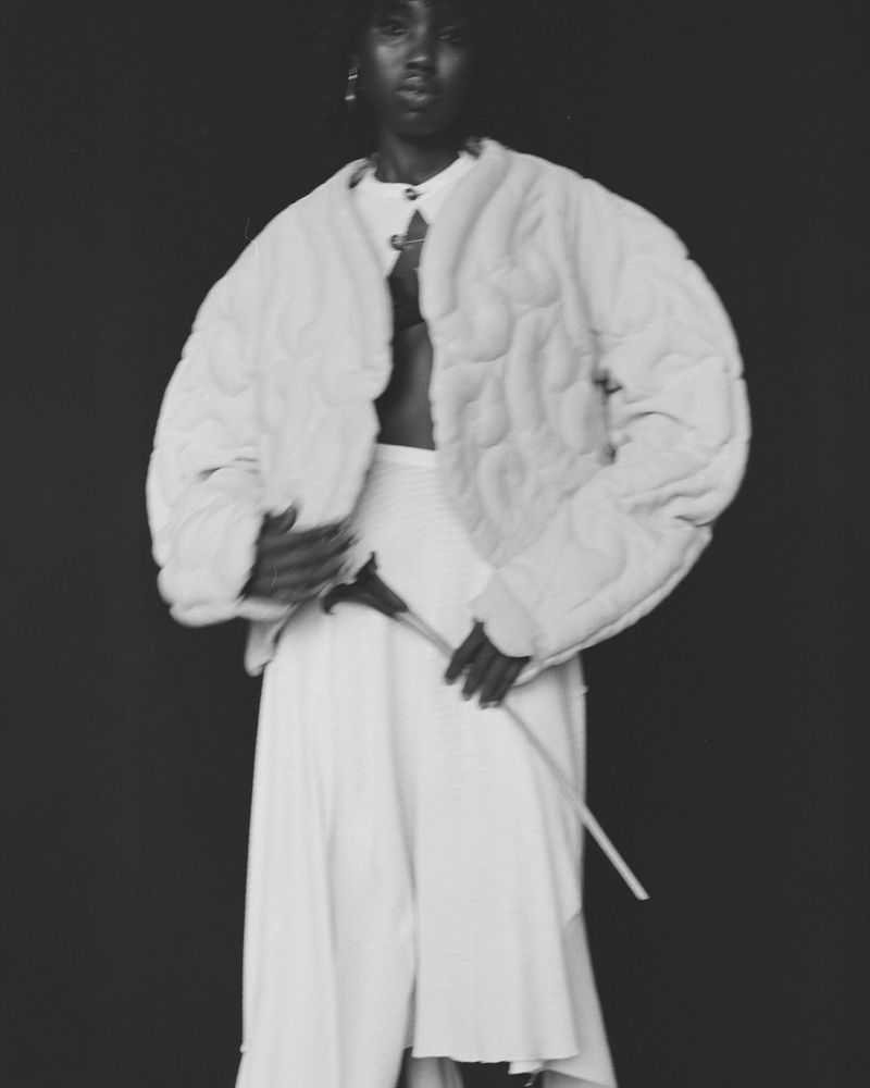 Fashionable African woman posing in a white coat and skirt against a dark background, holding a flower stem, in a studio setting.