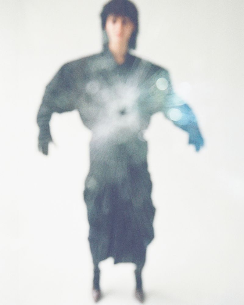 Unfocused studio shot of a person in a black outfit with dramatic shoulder silhouette, soft lighting, and a blurred bokeh overlay.