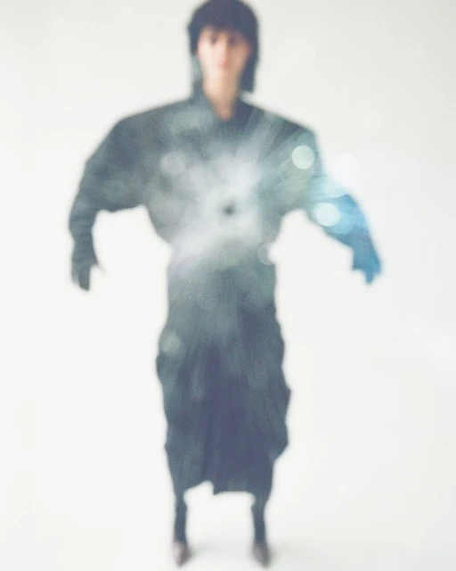 Unfocused studio shot of a person in a black outfit with dramatic shoulder silhouette, soft lighting, and a blurred bokeh overlay.