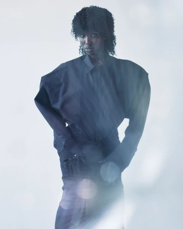 Model with textured curly hair wearing an oversized dark structural shirt with exaggerated shoulders against a bright background.
