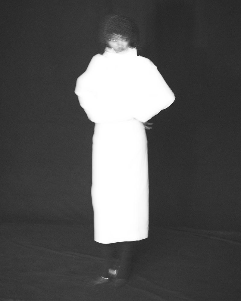 Model posing in a glowing white high-collar dress against a dark black backdrop in a monochrome fashion portrait.