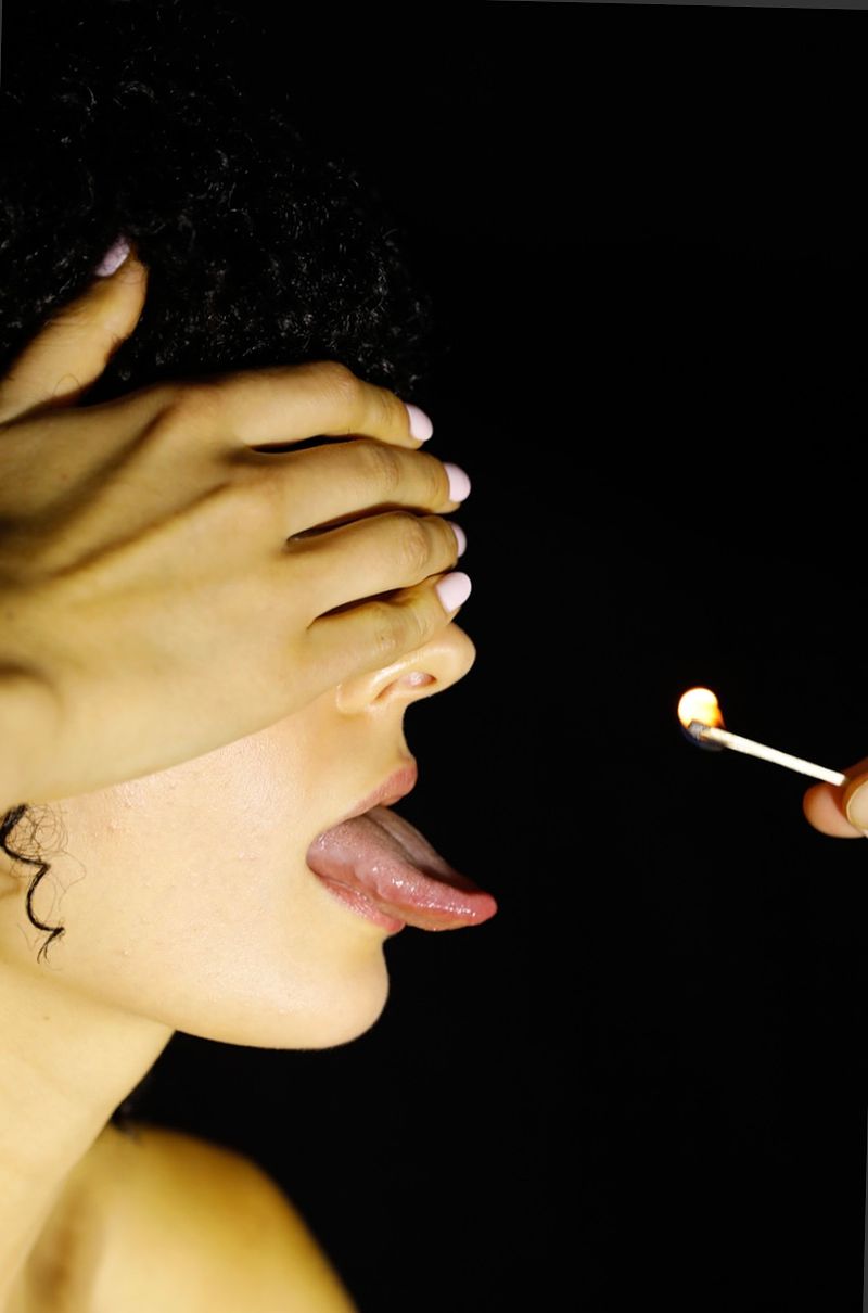 Close-up shot of a woman with her tongue out, eyes covered by her hand, facing a lit match, with a dark background and dramatic lighting.