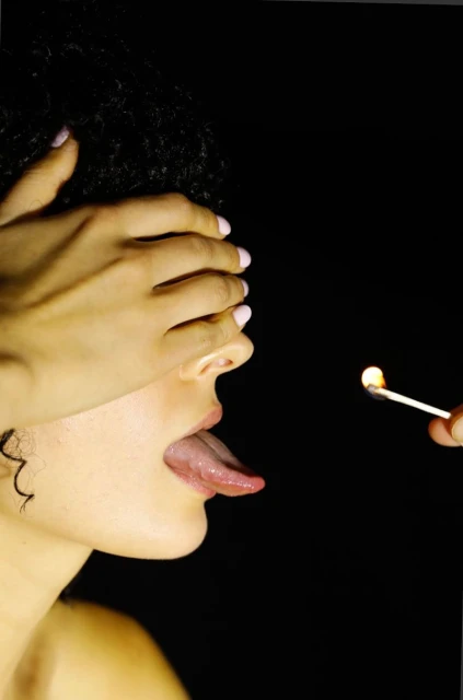 Close-up shot of a woman with her tongue out, eyes covered by her hand, facing a lit match, with a dark background and dramatic lighting.