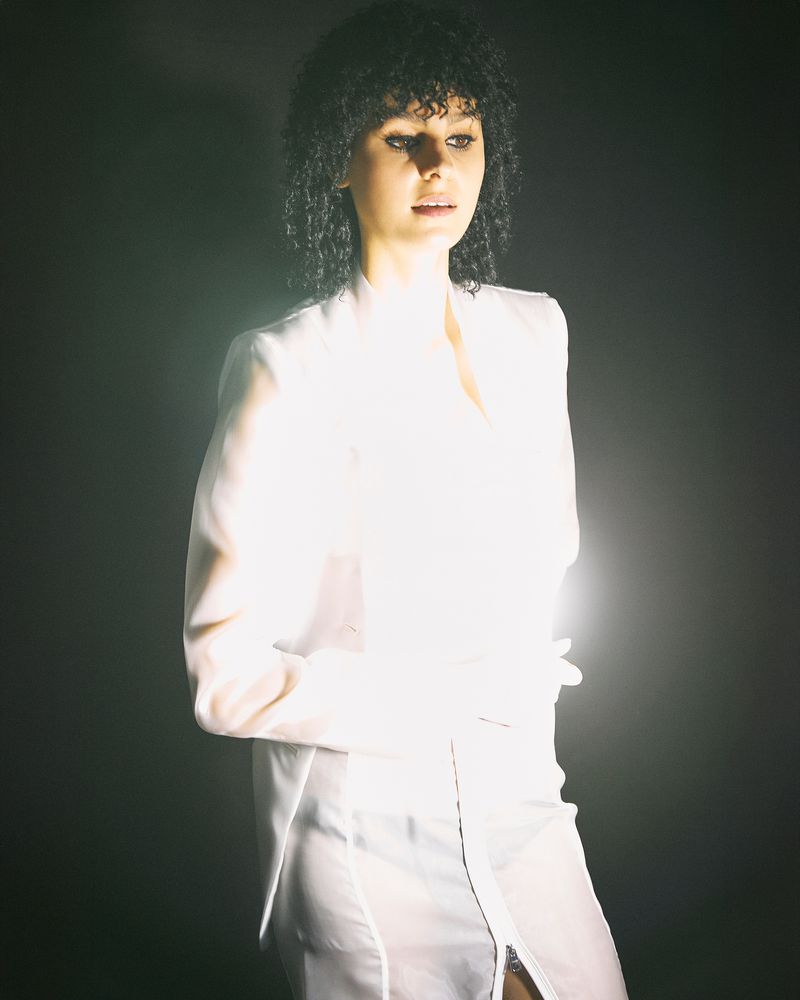 Portrait of a woman with curly hair wearing a white blazer in front of a dark backdrop with backlighting creating a bright halo effect.