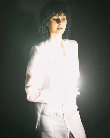 Portrait of a woman with curly hair wearing a white blazer in front of a dark backdrop with backlighting creating a bright halo effect.