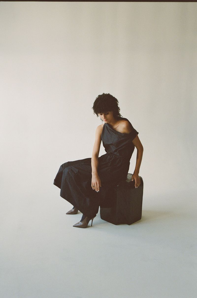 Fashionable adult woman in a black, one-shoulder dress and heeled boots, seated on a leather cube in a brightly lit studio.