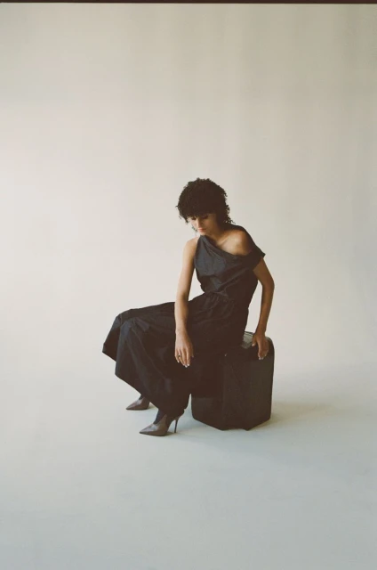 Fashionable adult woman in a black, one-shoulder dress and heeled boots, seated on a leather cube in a brightly lit studio.