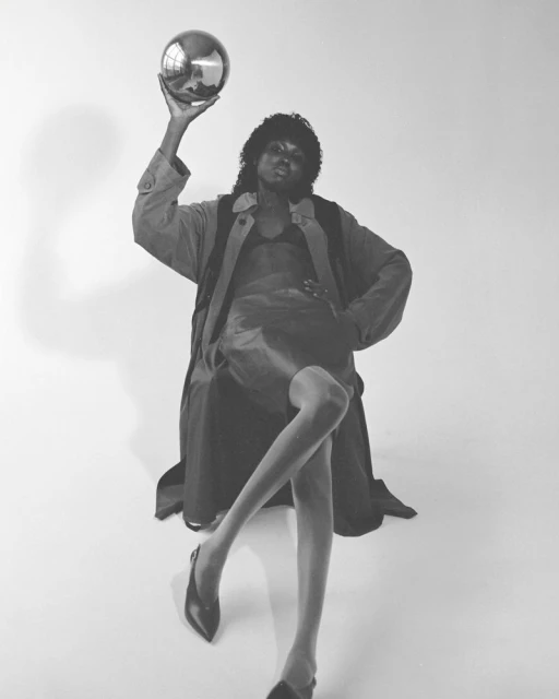 Elegant Black woman in stylish coat poses gracefully with a silver ball in a high-fashion photo studio setting with bright lighting.