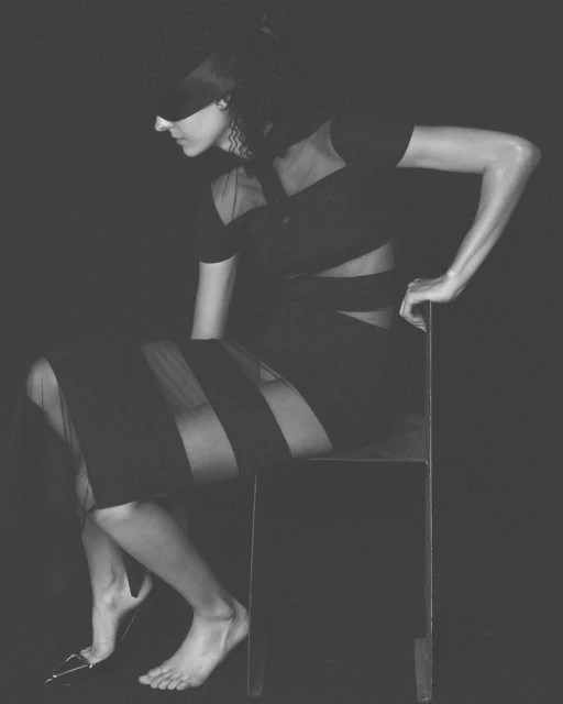 Monochrome studio portrait of a woman sitting on a chair wearing a black dress and blindfold in a dark, moody environment.