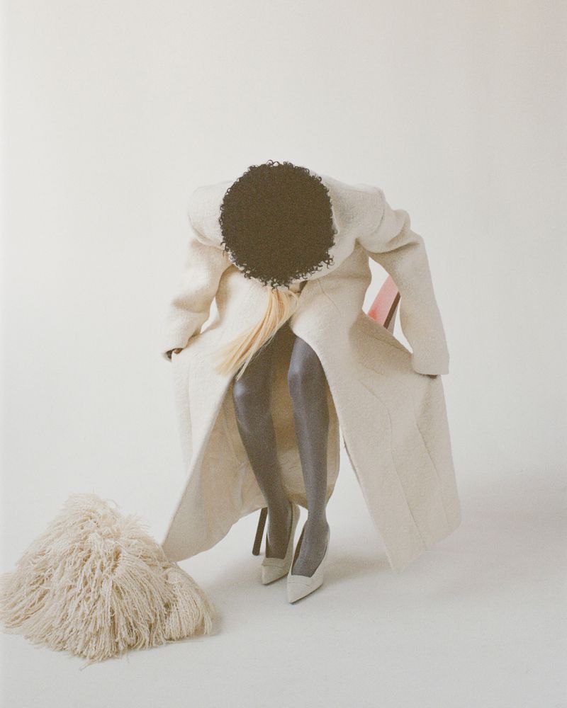 A woman in a long white coat sits on a chair, posing in high heels with a white shag rug in front of a plain white background.