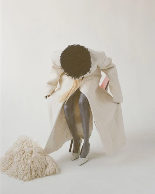 A woman in a long white coat sits on a chair, posing in high heels with a white shag rug in front of a plain white background.