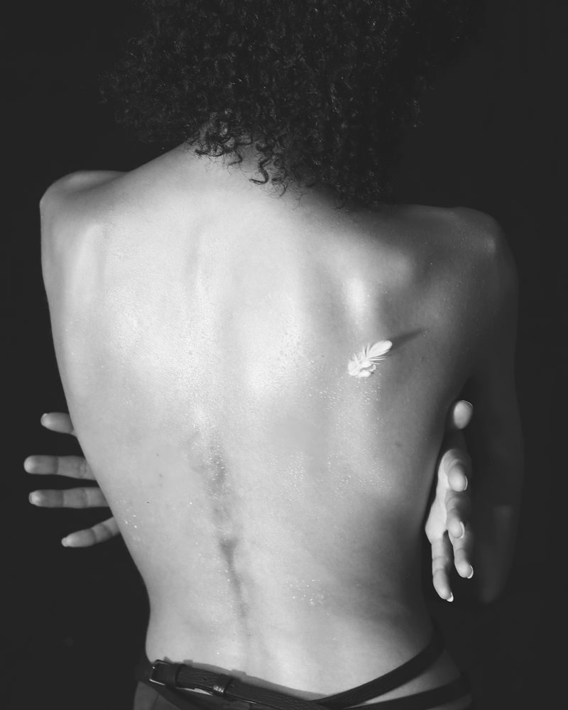 Black and white portrait focusing on the bare back of a person with a single white feather resting against the shoulder blade.