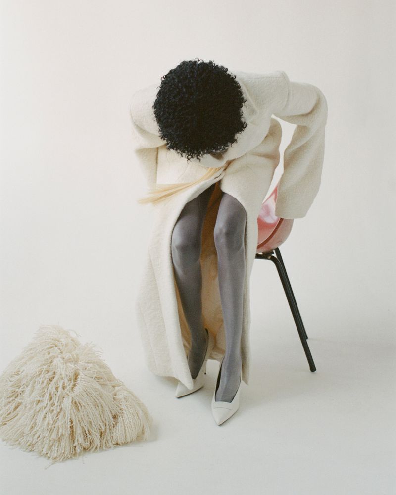 An African American woman in a white dress and gray stockings is sitting on a red chair in a white photo studio, and a white fur rug is on the floor.