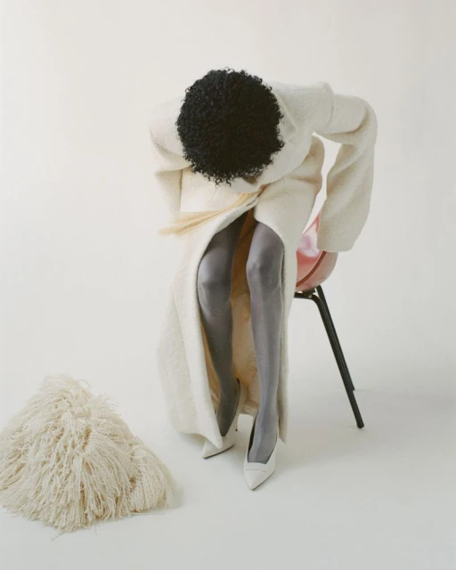 An African American woman in a white dress and gray stockings is sitting on a red chair in a white photo studio, and a white fur rug is on the floor.