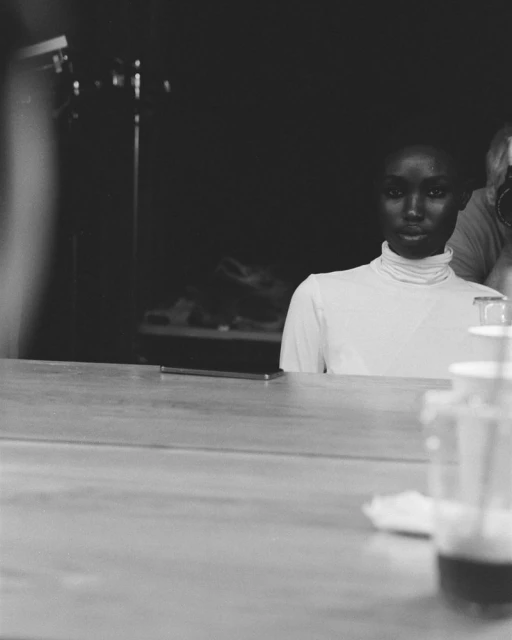 Monochrome close-up of a woman with a white turtleneck, a photographer, and a glass on a counter.
