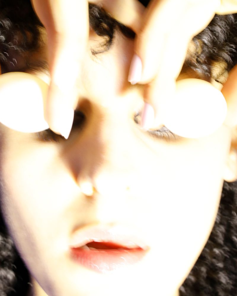 A close-up studio shot features a woman's face, with two hands holding white balls above her eyes, obscuring her forehead and hair.