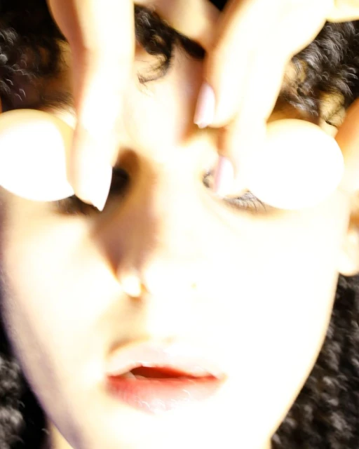 A close-up studio shot features a woman's face, with two hands holding white balls above her eyes, obscuring her forehead and hair.
