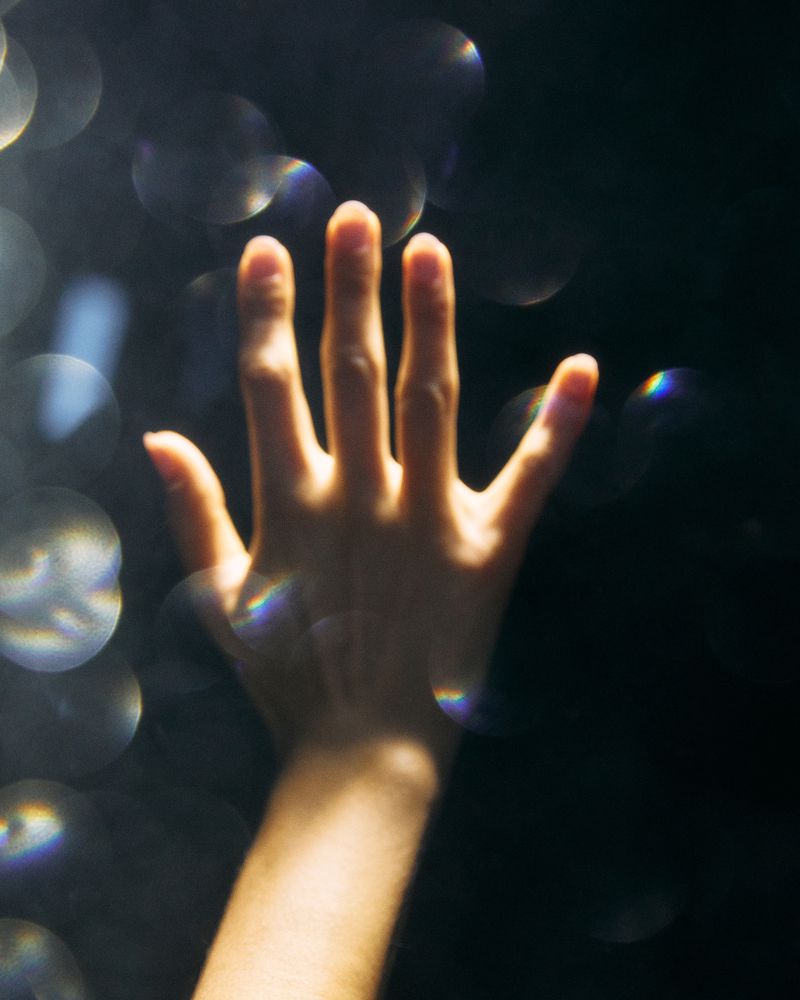Close-up of a human hand against a dark backdrop with bokeh and spectral highlights, creating a sense of ethereal reach.