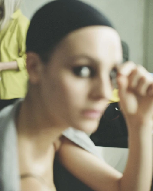 A woman wearing a wig cap is posing in a blurred studio environment, with another woman in a yellow shirt in the background.