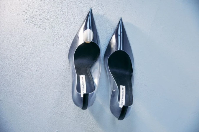 Elevated view of clear stiletto heels with pointed toes and a designer label set against a textured off-white background.