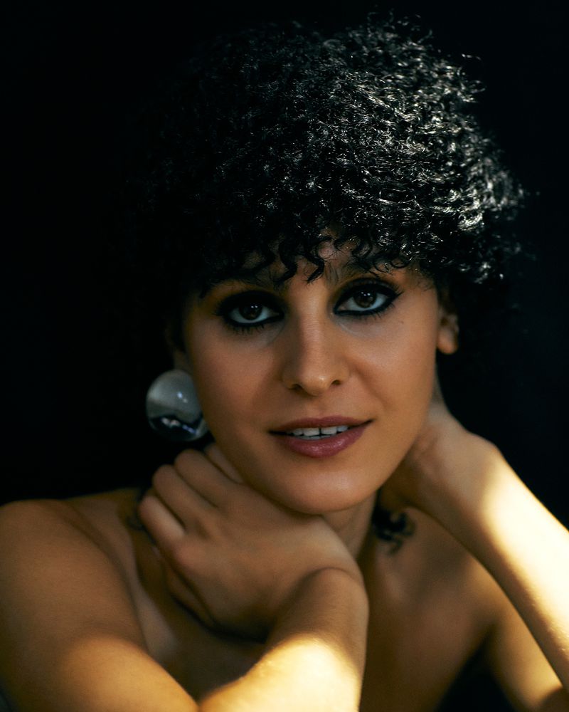 Close-up beauty portrait of a woman with short dark curly hair, smoky eye makeup, and a round earring against a dark background.