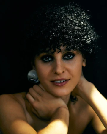 Close-up beauty portrait of a woman with short dark curly hair, smoky eye makeup, and a round earring against a dark background.