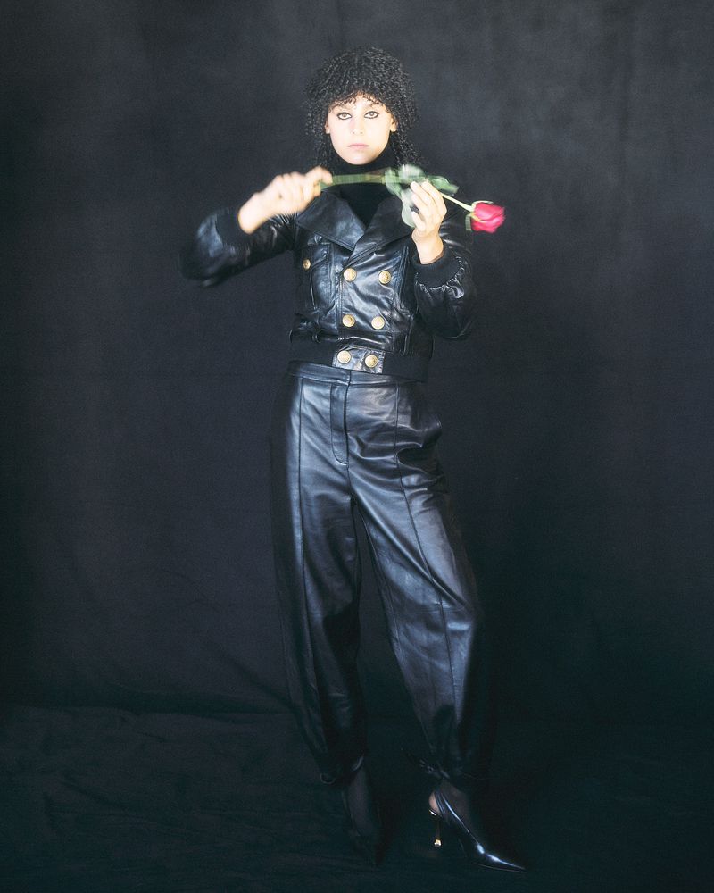A woman with dark curly hair in black leather jacket and pants, holding a red rose in a dramatic pose against a dark background.