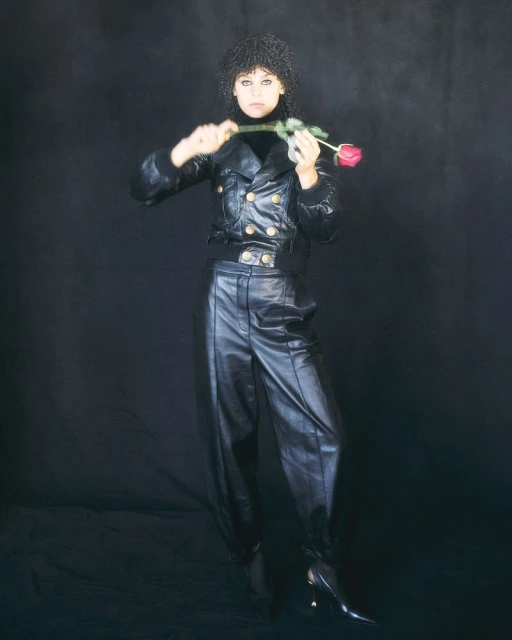 A woman with dark curly hair in black leather jacket and pants, holding a red rose in a dramatic pose against a dark background.