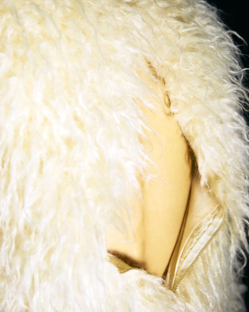 Close-up on a fluffy, cream-colored faux fur coat with a gold zipper. The jacket is photographed against a dark background.