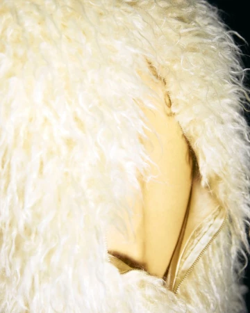 Close-up on a fluffy, cream-colored faux fur coat with a gold zipper. The jacket is photographed against a dark background.