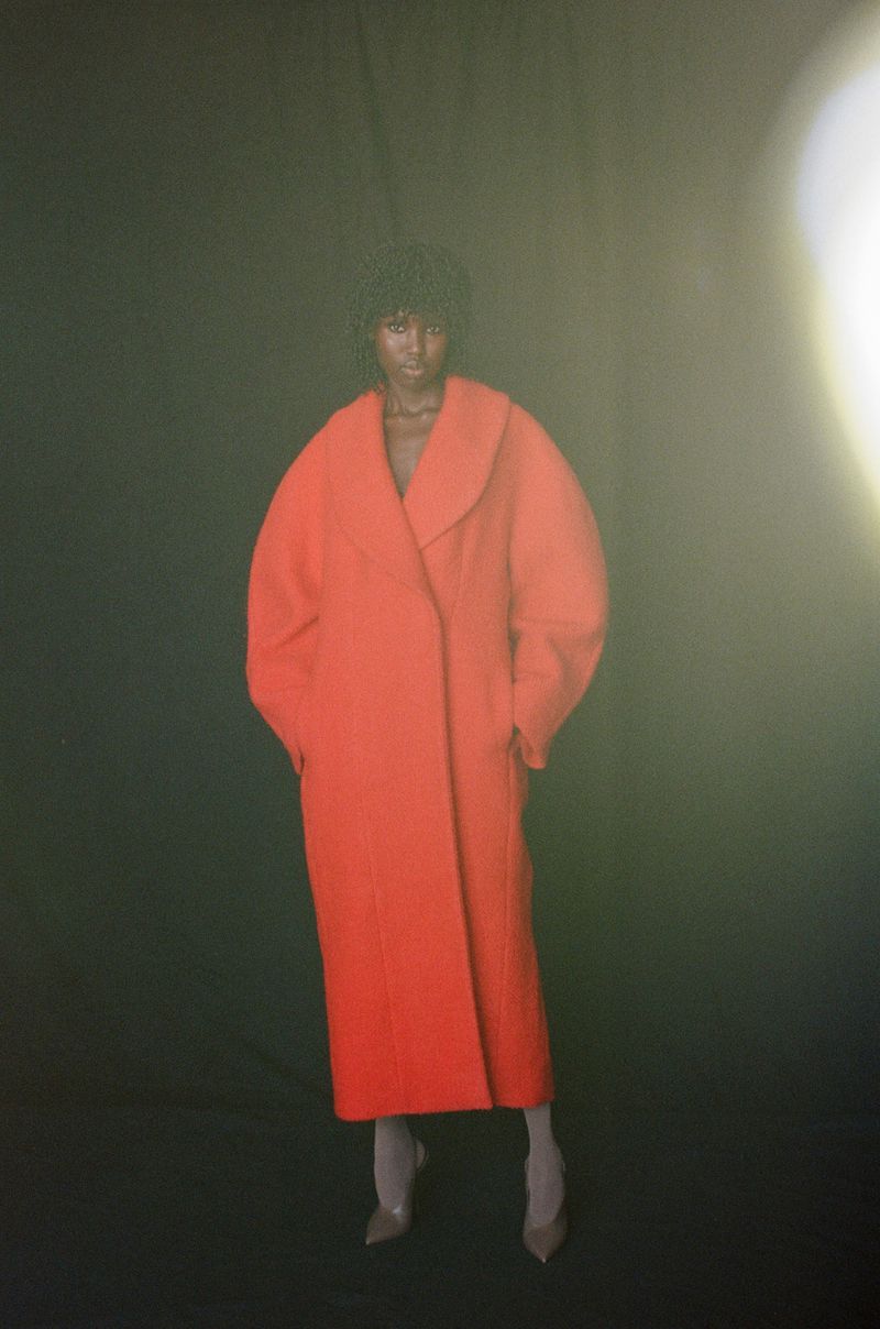 An adult Black female fashion model in a vibrant red oversized coat stands against a dark backdrop in a studio environment with soft lighting.
