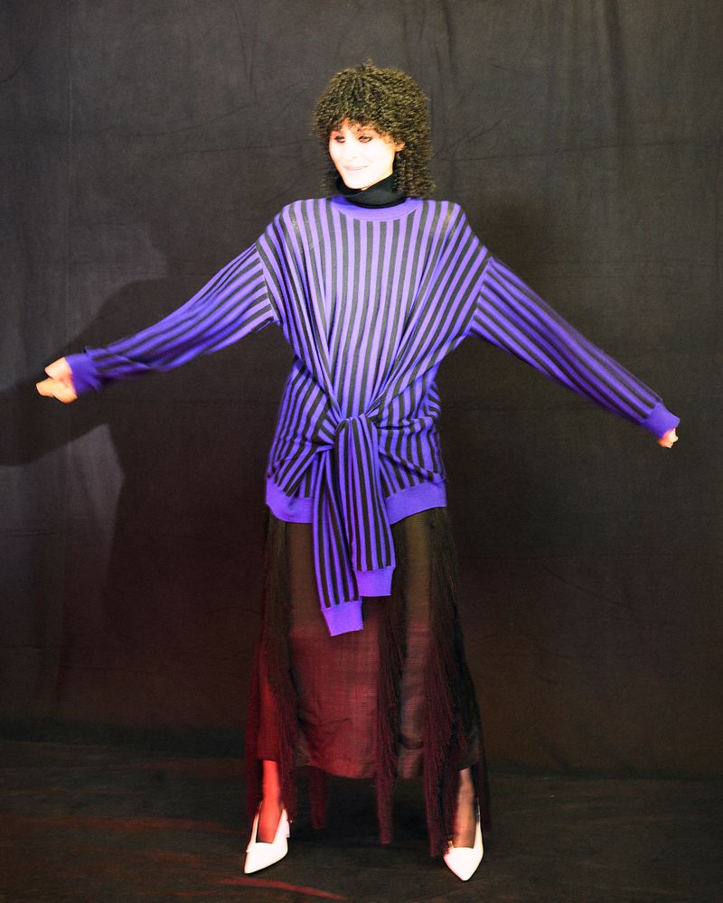 Woman in purple-and-black striped sweater and black skirt poses in a studio. She has dark curly hair, white heels, and a dark background.