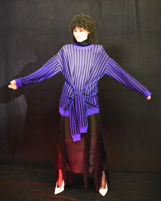 Woman in purple-and-black striped sweater and black skirt poses in a studio. She has dark curly hair, white heels, and a dark background.
