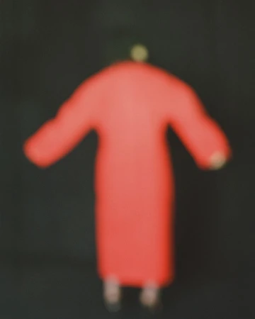 Heavily blurred silhouette of a model wearing a long vibrant red coat against a solid black background.