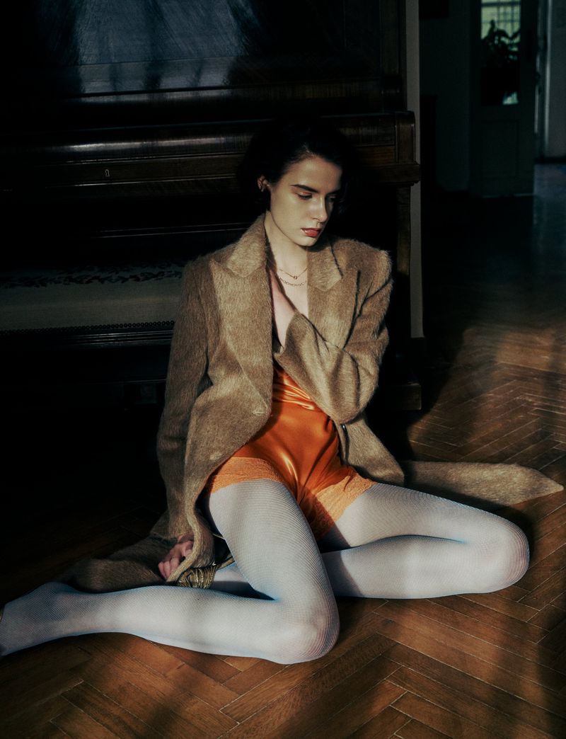 Young woman in an orange bodysuit, tights, and coat sits on a herringbone wood floor with a dark piano and muted light.