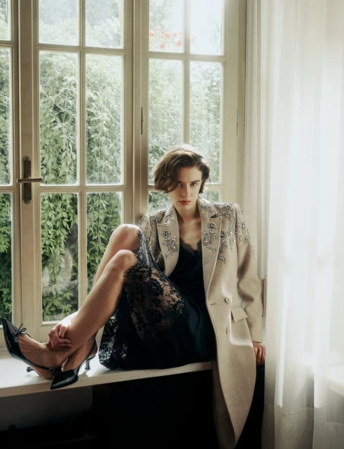 An adult woman sits by a window wearing an embellished coat, black lace dress, and black heels in an indoor, natural light setting.