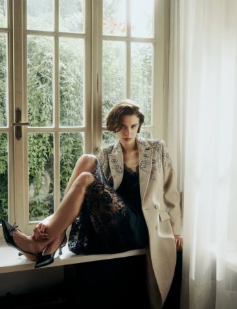 An adult woman sits by a window wearing an embellished coat, black lace dress, and black heels in an indoor, natural light setting.